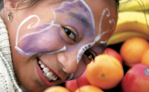 A happy child wearing face-paint grinning in front of a basket of popular fruits